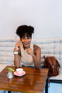 Delighted young african american female with dark curly hair in stylish clothes smiling while having phone conversation sitting in cafe at table with cup of coffee