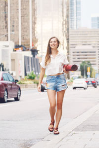 Full length of young woman carrying exercise mat while walking on footpath in city