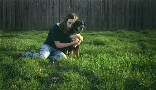 Young woman embracing dog while resting on grassy field in yard