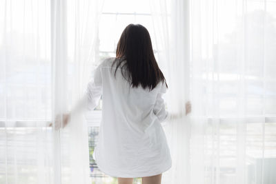 Rear view of woman looking through window by white curtains at home