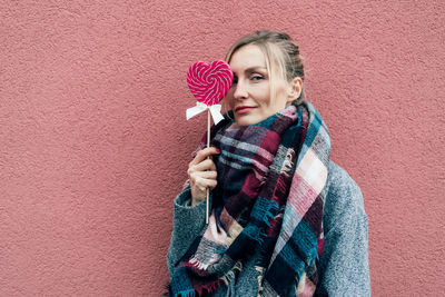 Portrait of woman holding heart shaped lollipop against wall