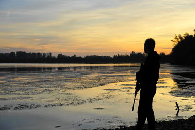 Silhouette man with rifle standing by lake during sunset