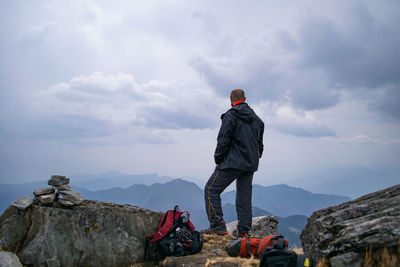 Rear view of mid adult man with backpacks standing on mountain against cloudy sky