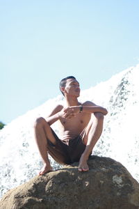 Full length of man sitting on rock against waterfall and sky