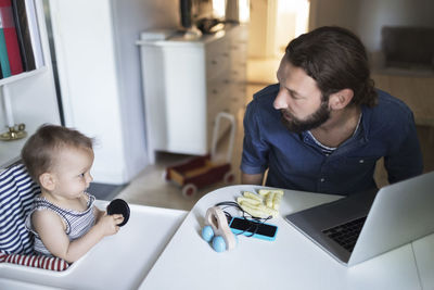 Mid adult man taking care of baby boy with laptop on table at home