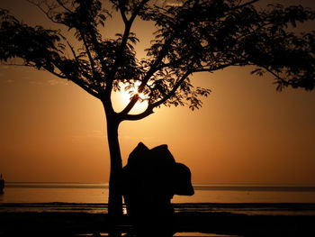Silhouette tree and sculpture by beach at sunset