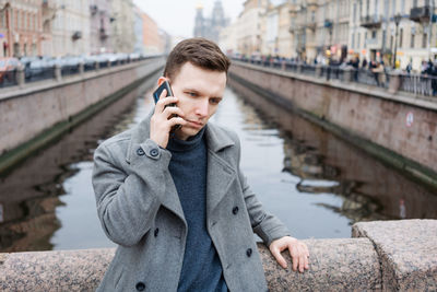 Emotional young man using mobile phone, wearing an elegant gray coat, stands