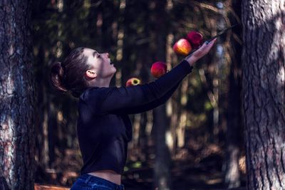 Side view of woman holding apples while standing by tree trunk