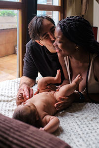 Loving hispanic man kissing cheek of smiling gentle african american mother while caressing cute infant baby lying on blanket