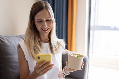 Smiling young woman using smart phone while holding food at home