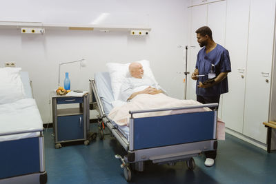 Male nurse talking to senior patient lying on bed at hospital