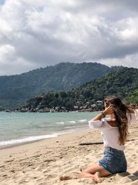 Rear view of young woman sitting at sandy beach against cloudy sky