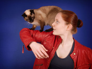 Young woman with siamese cat on arm against blue background