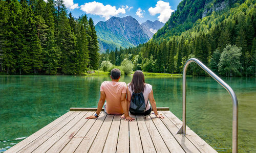 Rear view of young couple sitting on deck by lake in mountains.
