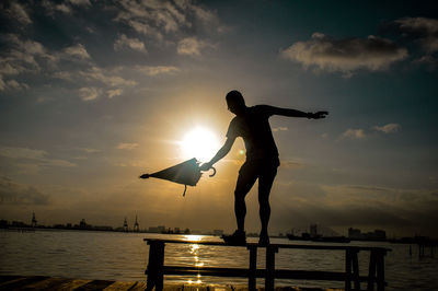 Silhouette man with umbrella balancing on bench against lake during sunset