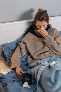 Sick young woman holding coffee cup while resting on bed