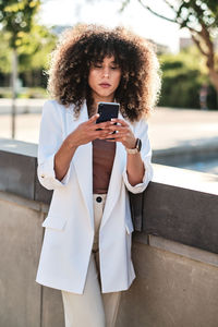Hispanic female entrepreneur in white suit with curly hair browsing on smartphone while standing near fence on sunny day on city street