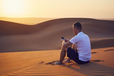 Alone in desert. young man sitting on sand dune and using phone at golden sunset.