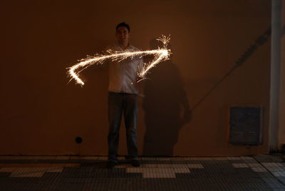 Full length portrait of man holding illuminated sparkler against wall at night