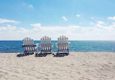 Empty adirondack chair on sand at beach against sky