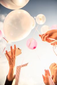 Low angle view of hands holding helium balloons against sky