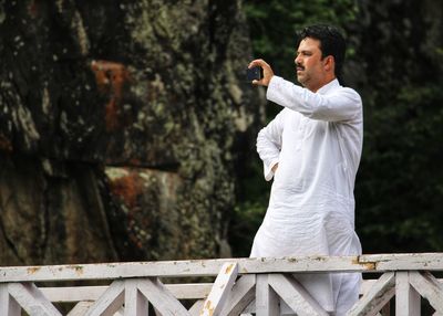 Young man photographing with smart phone while standing by railing against trees