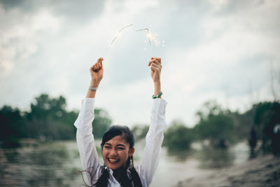 Cheerful young woman holding illuminated sparklers by lake against sky