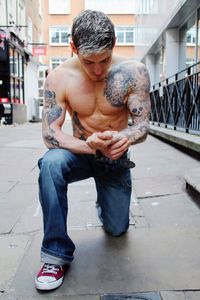 Shirtless tattooed man kneeling on footpath 