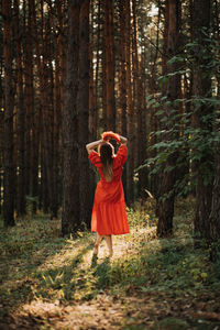 Barefoot happy young woman in red dress with hand raised dancing in pine forest at summer day.