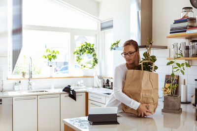 Woman holding paper bag while looking at digital tablet in kitchen