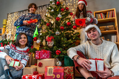 Group of four young friends dressed in christmas sweatshirts give gifts under the christmas tree