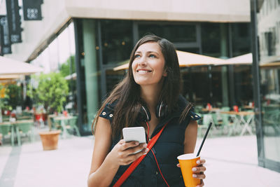 Portrait of smiling young woman with cell phone and takeaway drink in the city