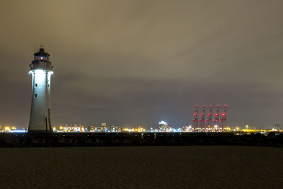 Illuminated lighthouse by building against sky at night