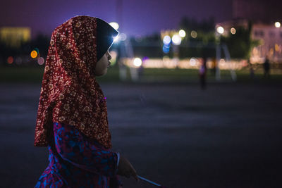 Side view of girl wearing hijab in illuminated city at night