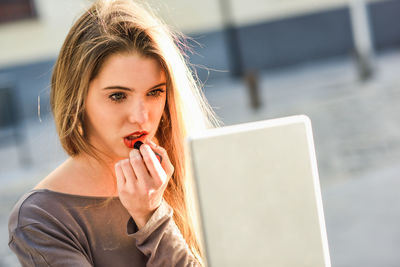 Beautiful young woman applying lipstick while looking at digital tablet