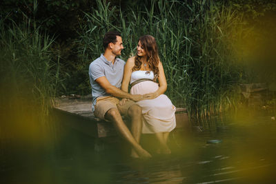 Man kissing pregnant wife while sitting on pier over lake