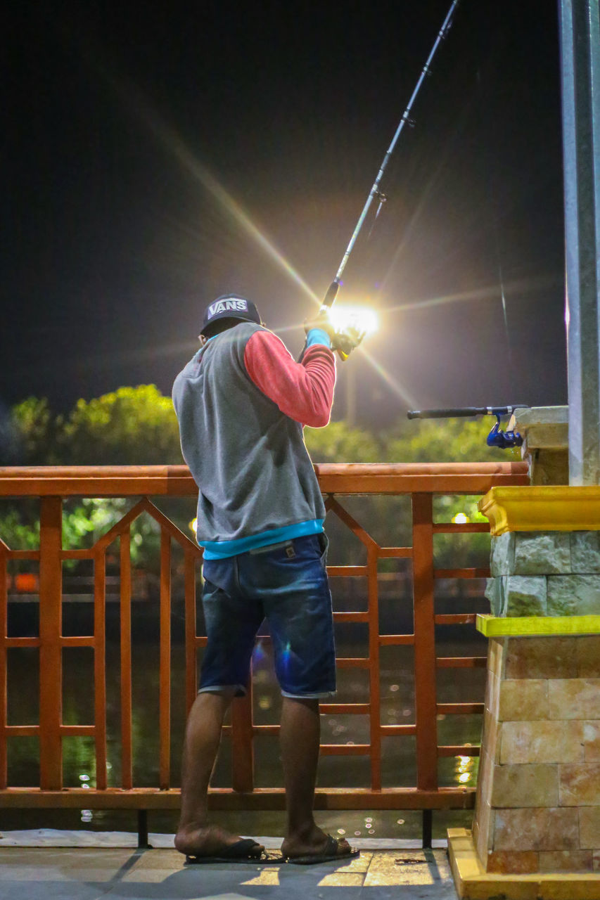 REAR VIEW OF MAN STANDING BY RAILING AGAINST ILLUMINATED LIGHTS