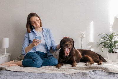 Smiling woman using phone while sitting with dog on bed