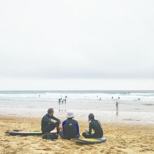 People with surfboards relaxing at beach against cloudy sky