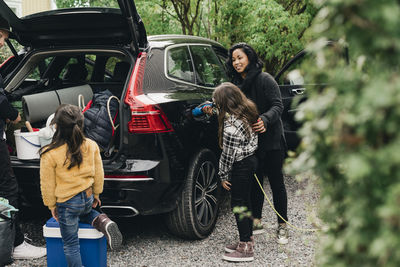 Woman guiding daughter in charging electric car while family loading luggage