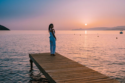 Woman standing on pier by sea against sky during sunset