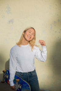 Smiling young woman with skateboard standing against wall