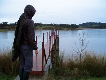 Rear view of man in hooded jacket standing by pier over lake