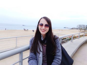 Portrait of smiling young woman standing by railing at promenade against sea