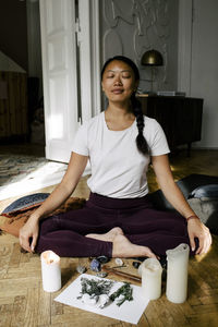 Smiling young woman meditating in front of herbs and lit candles at home