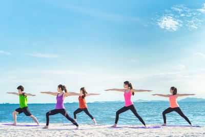 Young friends practicing yoga in warrior position on shore at beach during sunny day