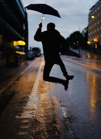 Rear view of man holding umbrella jumping on wet street at dusk