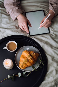 Woman using digital tablet for drawing while lying on bed and enjoying croissant with cappuccino	
