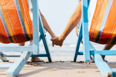 Low section of couple holding hands sitting on chair at beach