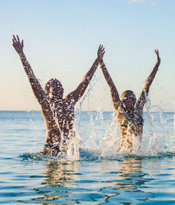 Portrait of two people emerging from underwater with hands raised high 